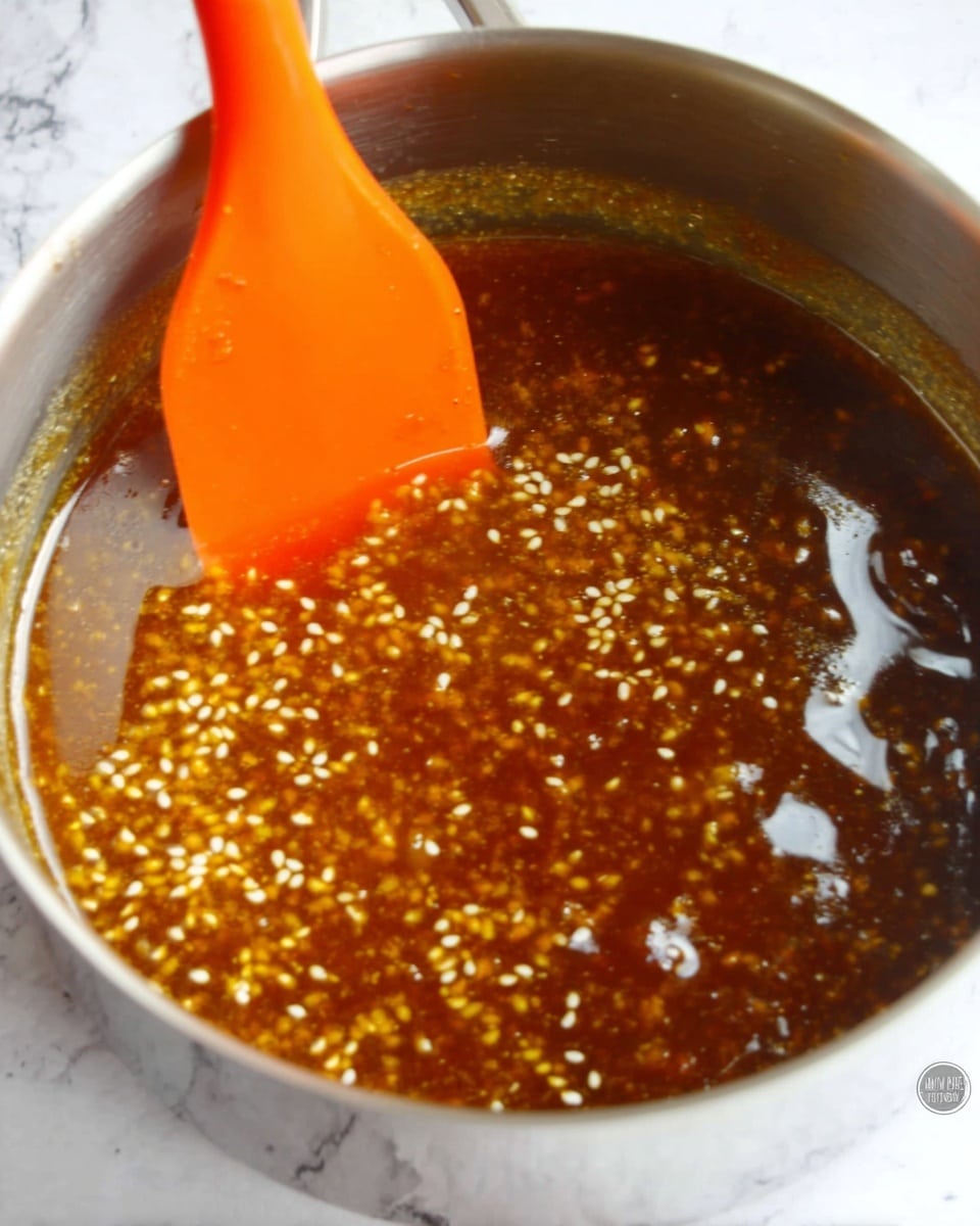 The image shows a close-up of a silver pot filled with a shiny golden brown liquid sauce mixed with small white sesame seeds floating on the surface. An orange spatula is partially inside the pot, resting against the side, with drops of sauce on it. The background is a white marbled texture. photo taken with an iphone --ar 4:5 --v 7