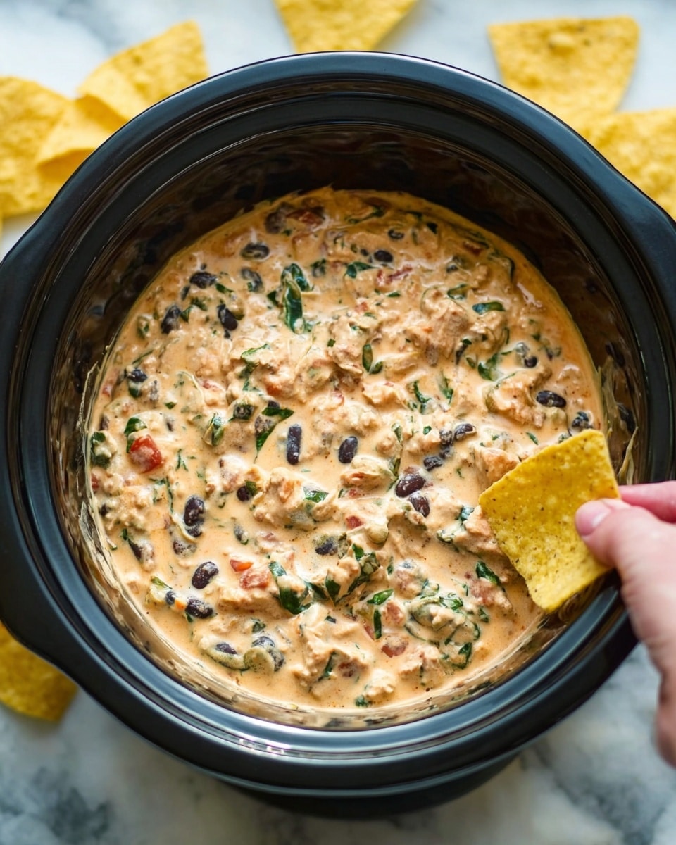 The image shows a black slow cooker filled with a creamy dip that has a mix of colors and textures. The top layer is thick and creamy with visible chunks of light brown meat, dark black beans, and small bits of green spinach or herbs, all mixed together in a light orange sauce with some flecks of red. The dip looks smooth but chunky at the same time, evenly spread inside the cooker. Around the cooker, on a white marbled surface, there are yellow tortilla chips that add a crunchy contrast to the creamy dip. A woman's hand, holding a chip, is reaching towards the slow cooker from the side. Photo taken with an iphone --ar 4:5 --v 7