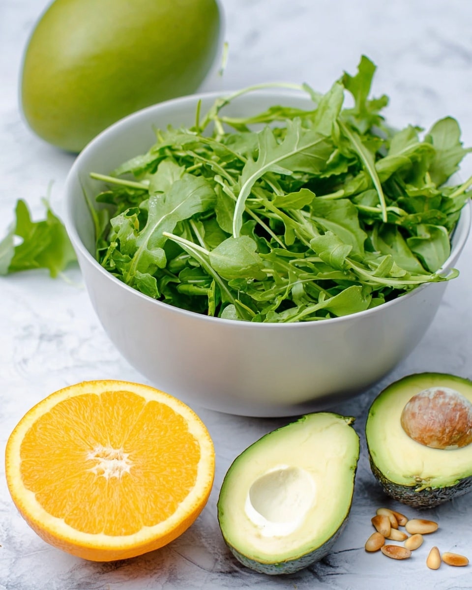 A white bowl filled with fresh green arugula leaves, showing different shades and textures of leafy greens, sits on a white marbled surface. In front of the bowl, there is a half orange with bright orange flesh and white center, two halves of a ripe avocado with creamy light green flesh and one half showing the large brown pit. A few small pine nuts are scattered near the avocado. In the background, a whole green mango is partially visible. The colors are bright and natural, with a fresh and healthy look. photo taken with an iphone --ar 4:5 --v 7