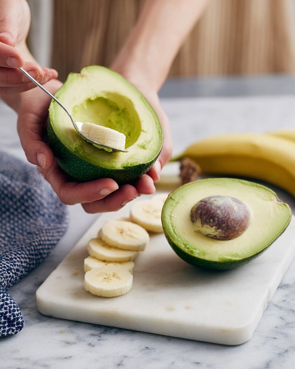 The image shows a close-up of a woman's hands holding a halved avocado with bright green flesh and a smooth, creamy texture. One half of the avocado has the large brown seed still inside, while the other half is being scooped out with a small silver spoon. Below the avocado halves, there are several banana slices, pale yellow with a soft texture, placed on a white marbled cutting board. To the side, there is a blue and white patterned cloth on the white marbled surface. The background is softly blurred with light, neutral colors. photo taken with an iphone --ar 4:5 --v 7