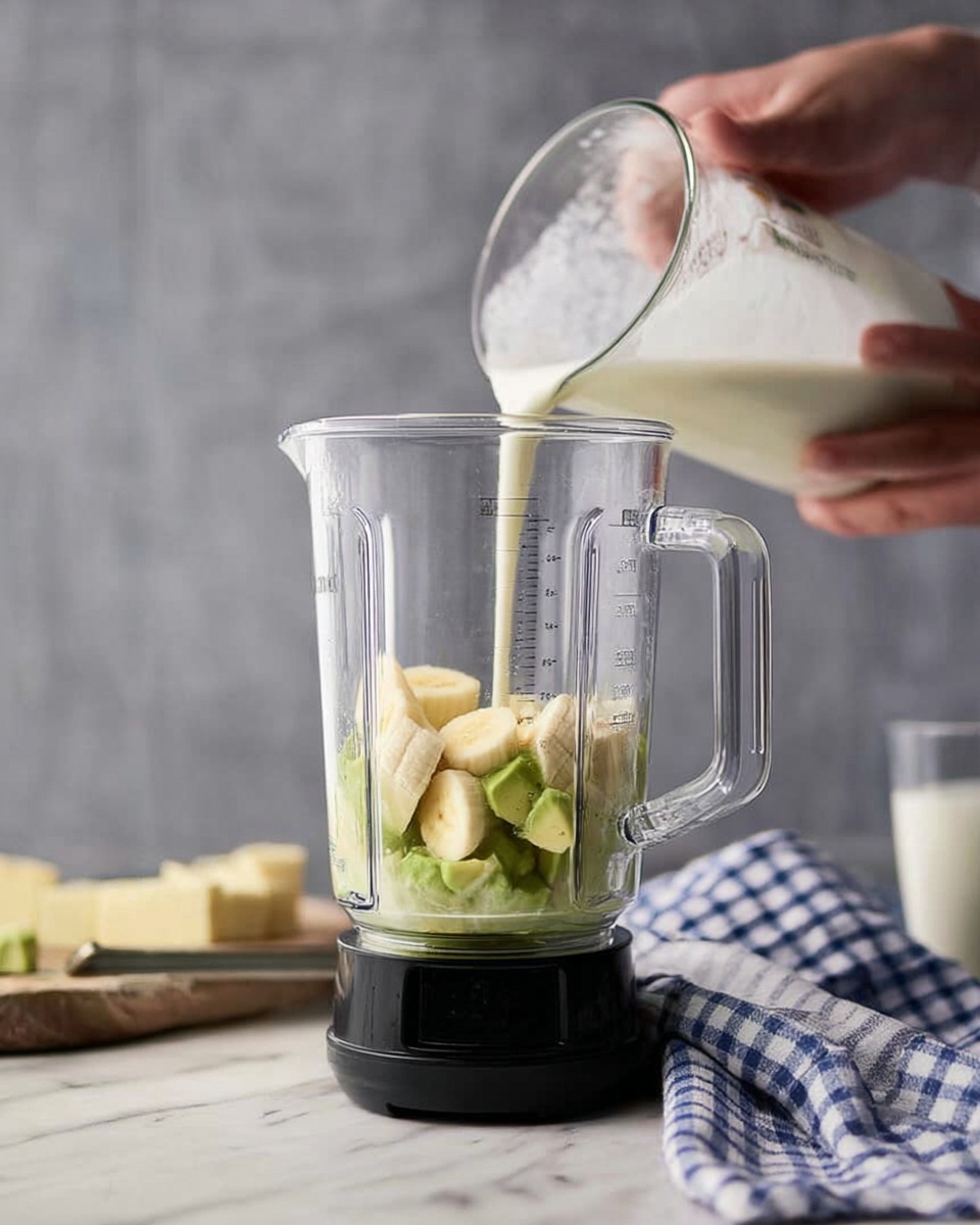 A clear blender jar is placed on a black base with chunks of light yellow banana and green avocado inside. A woman's hand is holding a clear measuring cup above the blender, pouring white milk into it. The background has a soft gray tone, and a white marbled surface holds a folded blue and white checkered cloth near the blender. Photo taken with an iphone --ar 4:5 --v 7