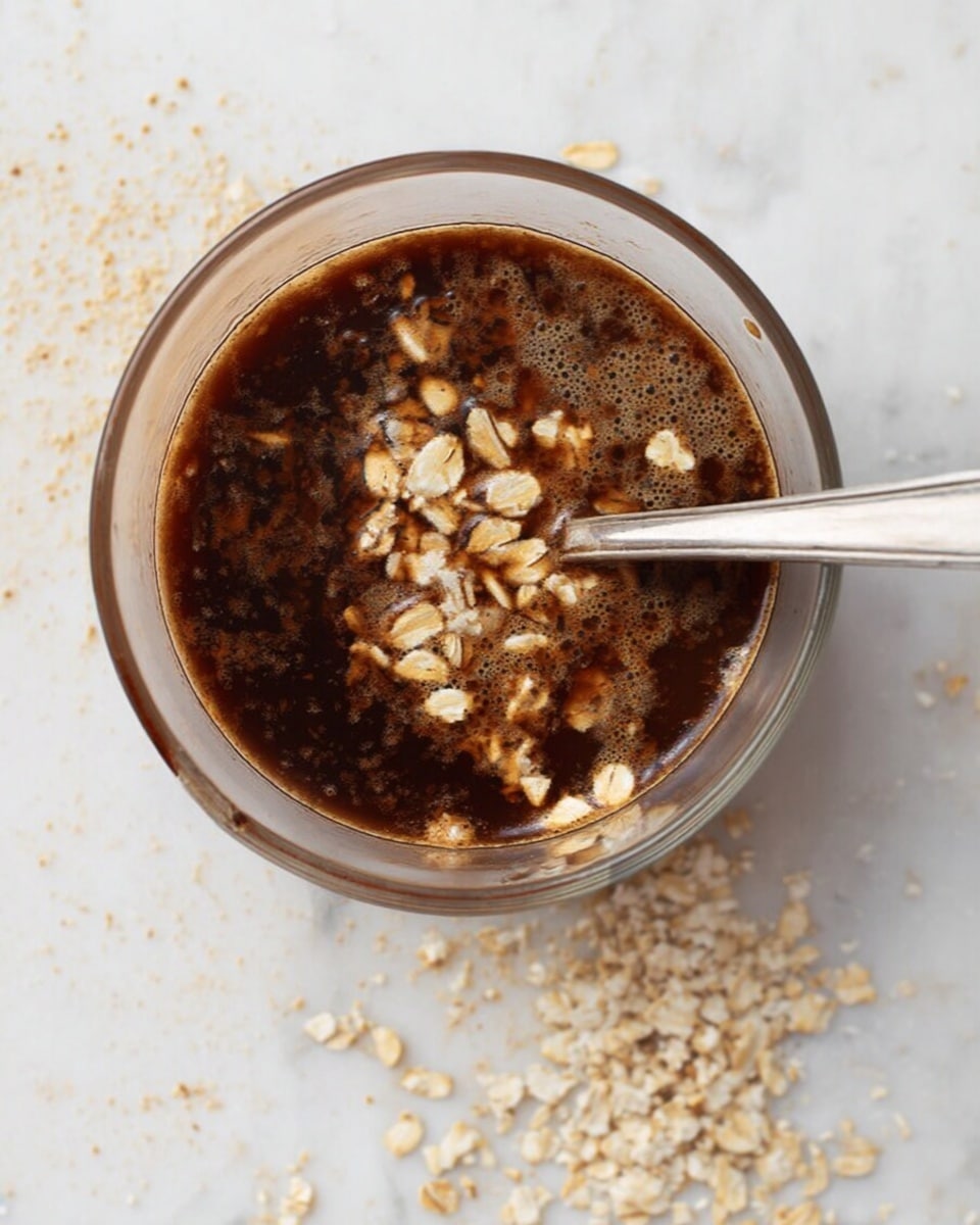 A clear glass cup filled with a dark brown, thick liquid mixed with light beige oat flakes floating on top. A silver spoon is placed inside the cup on the right side. Some oat flakes are scattered around the cup on a white marbled surface. The texture looks slightly grainy with visible bits of oats in the liquid photo taken with an iphone --ar 4:5 --v 7