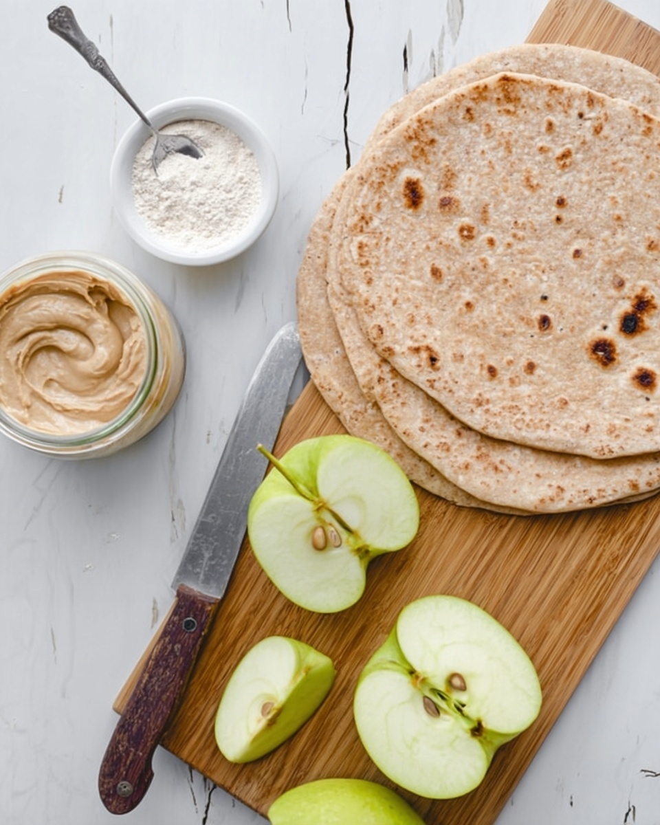 The image shows two light brown flatbreads stacked on top of each other with small darker brown spots scattered across their surface, placed on a white marbled texture. Below the flatbreads, there is a wooden cutting board with a silver knife with a dark wooden handle resting on it, alongside three green apple pieces: one whole quarter, one half apple with seeds visible, and one smaller slice. To the left of the cutting board, there is a small glass jar filled with a creamy beige spread and a silver spoon inside. Next to the jar, there is a small white bowl filled with a fine white powder. The overall scene is bright and clean. Photo taken with an iphone --ar 4:5 --v 7