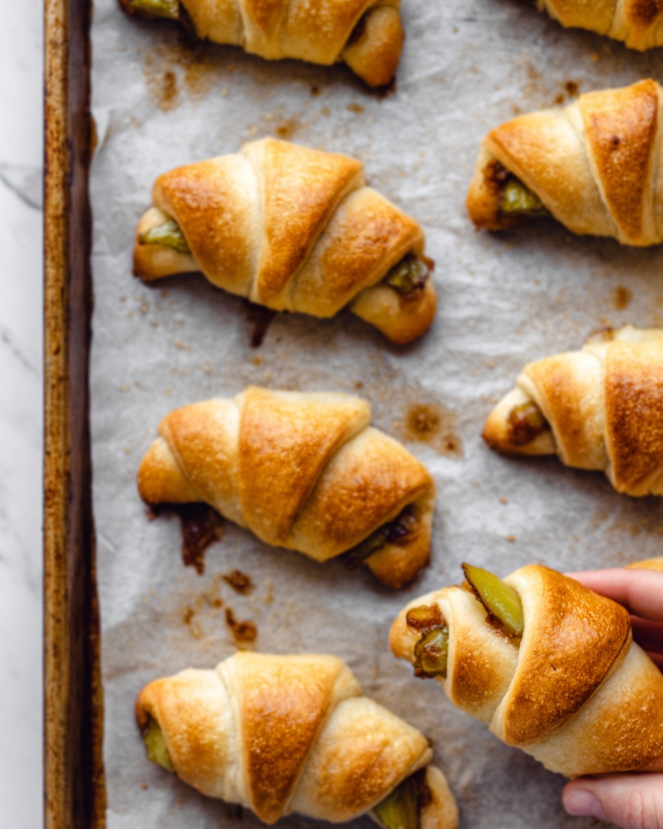 The image shows several golden-brown crescent rolls baked on a baking tray lined with parchment paper. Each roll is folded into a triangle shape with soft, flaky, and slightly crispy texture on the outer layer. Inside the rolls, slices of green pickles and some brown filling are visible, slightly peeking out from the ends. The parchment paper underneath has some shiny spots where oil or juices have leaked during baking, adding a subtle glisten to the scene. The rolls are arranged in rows on the tray, and a woman's hand is gently holding one roll on the right side of the image. The background is a white marbled texture. Photo taken with an iphone --ar 4:5 --v 7