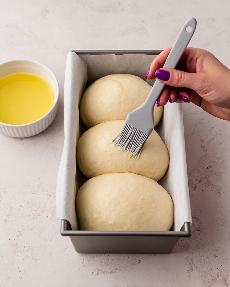 There are three large, smooth, pale dough balls sitting next to each other inside a loaf pan lined with white baking paper. A woman's hand with purple nails is holding a gray brush, gently spreading a liquid over the dough on the left side. Behind the pan, there is a white bowl with yellow liquid in it. The scene is set on a white marbled surface. Photo taken with an iphone --ar 4:5 --v 7