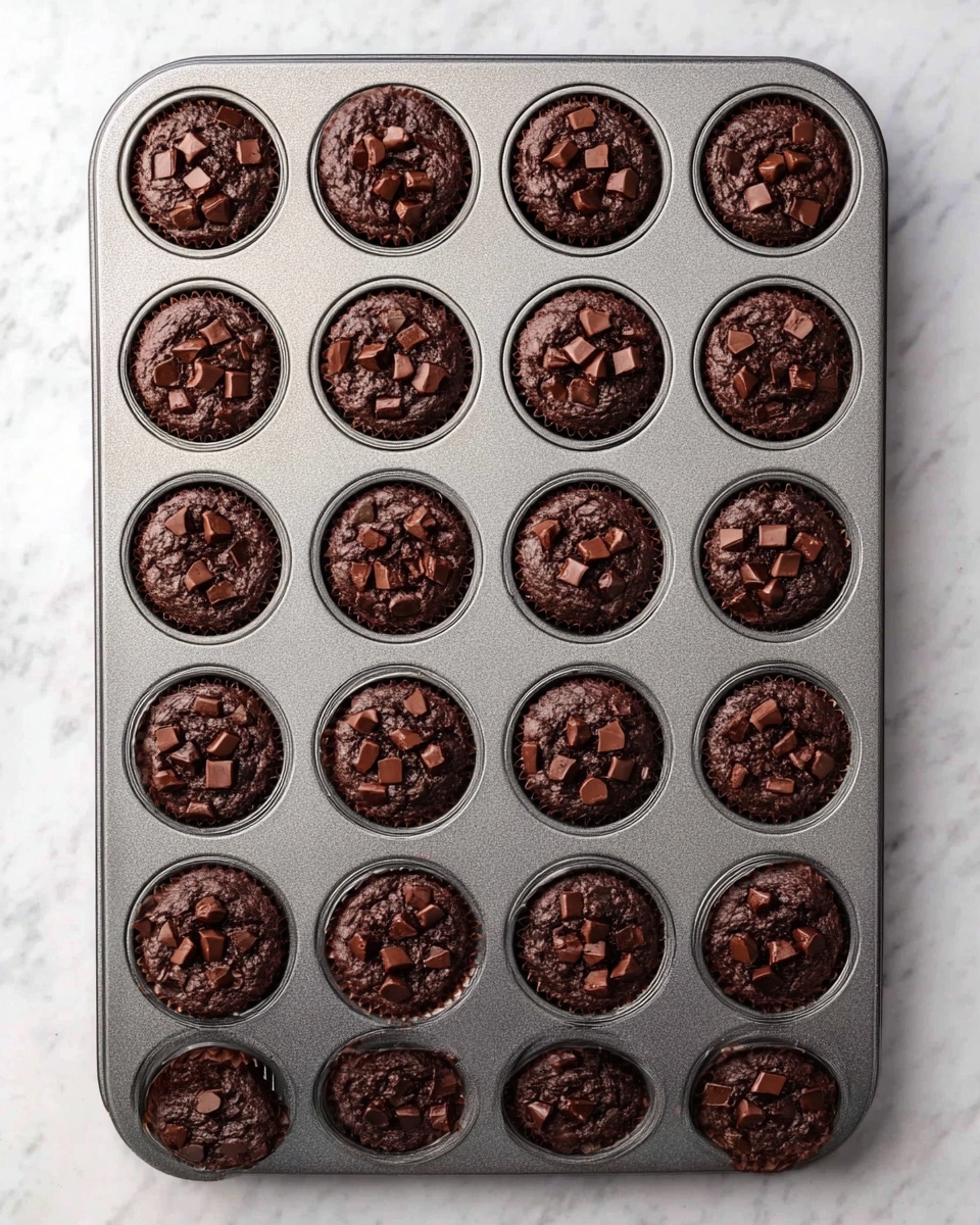 A gray metal muffin tray holds 24 freshly baked chocolate muffins arranged in a 4 by 6 grid. Each muffin is dark brown and topped with small chunks of chocolate, giving a rough texture on top. The muffins fit perfectly in the round cup holders. The tray sits on a white marbled surface, creating a clean and simple background. photo taken with an iphone --ar 4:5 --v 7