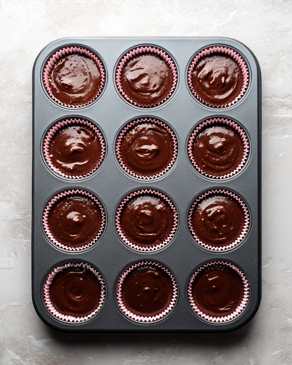 A dark gray metal baking tray with twelve round molds is placed on a white marbled surface. Each mold is lined with a light pink paper cupcake liner, and all liners are filled with a thick, smooth dark brown chocolate batter evenly spread inside. Small droplets and slight unevenness on the batter surface give a fresh and glossy look. The tray is seen from above with natural light softly highlighting the rich chocolate color. Photo taken with an iphone --ar 4:5 --v 7