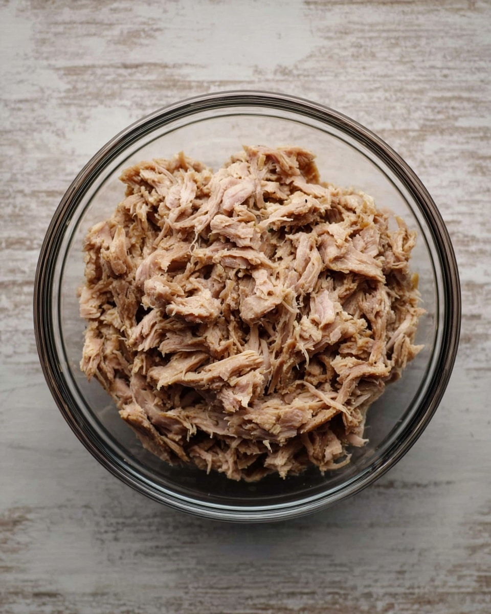 A clear glass bowl filled with shredded light brown meat sits on a white marbled texture surface. The meat is layered loosely, showing a tender and fibrous texture with some small chunks and strands visible. The bowl has a simple, smooth shape and the glass is transparent, allowing a clear view of the meat inside. Photo taken with an iphone --ar 4:5 --v 7