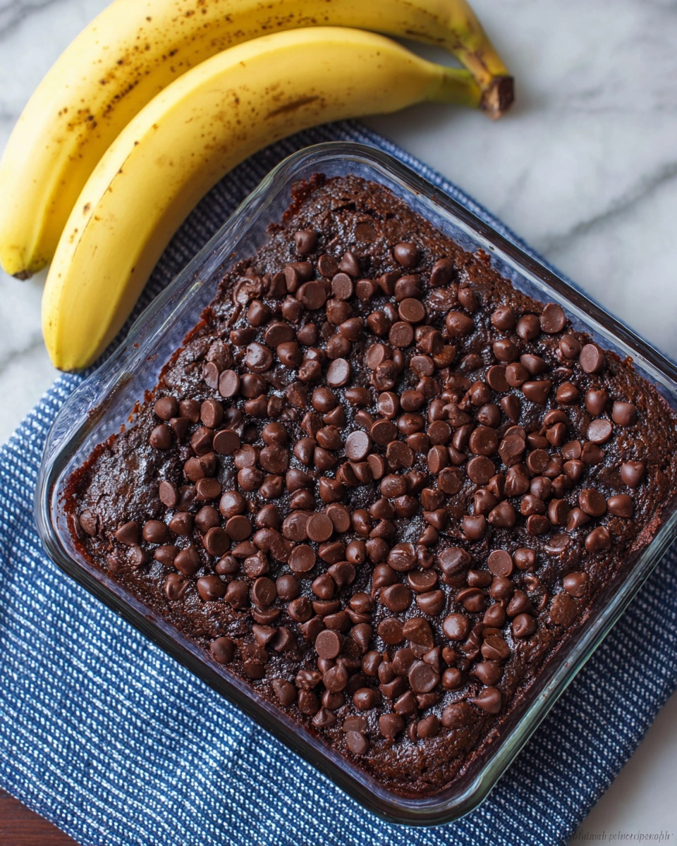 A square glass baking dish holds a single layer of dark brown chocolate banana bread topped thickly with many small, shiny, dark brown chocolate chips that cover the whole surface. The bread looks soft and moist with slightly uneven texture. The dish sits on a blue and white striped cloth on a white marbled surface. Next to the dish, there are two whole yellow bananas with small brown spots. Photo taken with an iphone --ar 4:5 --v 7