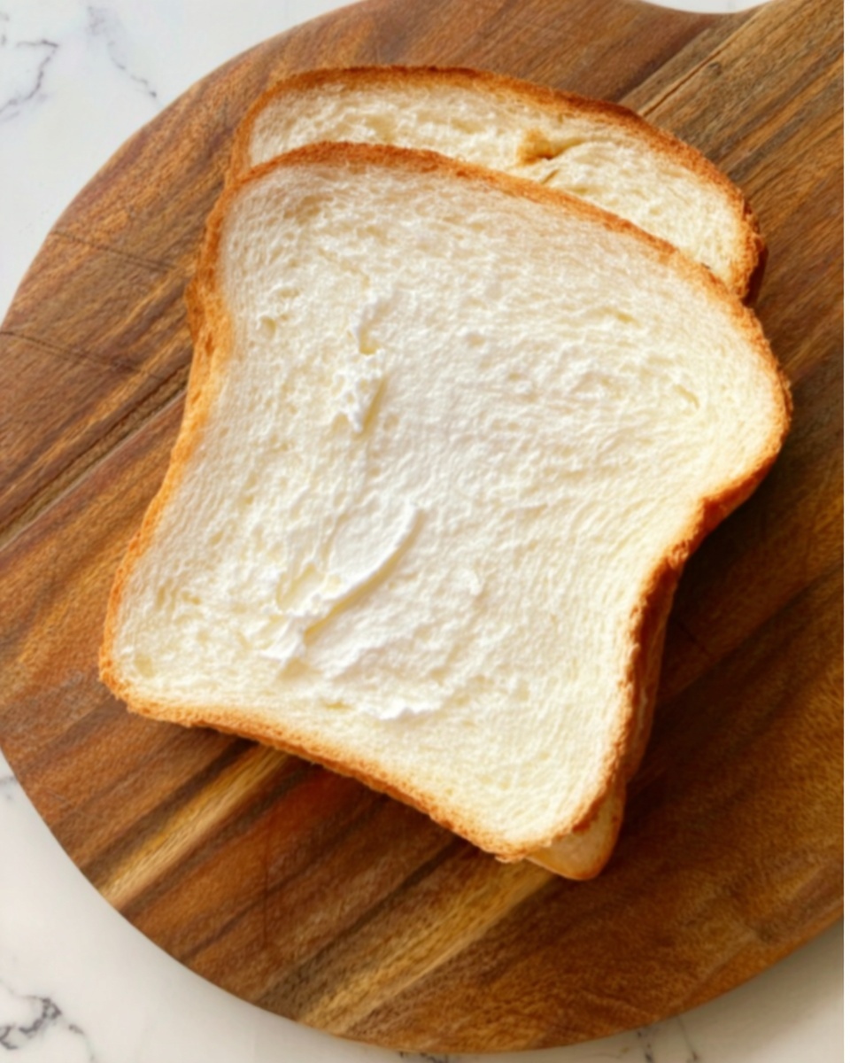 A single slice of soft white bread with a light airy texture is placed on a round wooden cutting board with a natural grain pattern. The board rests on a white marbled surface, enhancing the contrast between the bread's pale color and the wooden background. The slice shows slight indentations and a fluffy interior with a smooth crust around the edges. Photo taken with an iphone --ar 4:5 --v 7