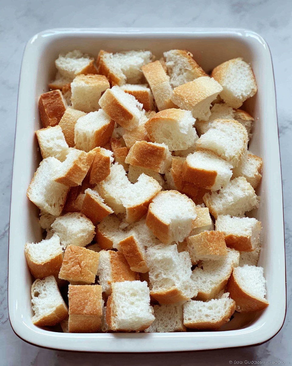 A rectangular white baking dish filled with many pieces of white bread cut into medium-sized cubes. The bread cubes show a soft, airy white interior with golden brown edges. The pieces are unevenly scattered inside the dish, filling it nearly to the top. The dish is placed on a surface with a white marbled texture. photo taken with an iphone --ar 4:5 --v 7