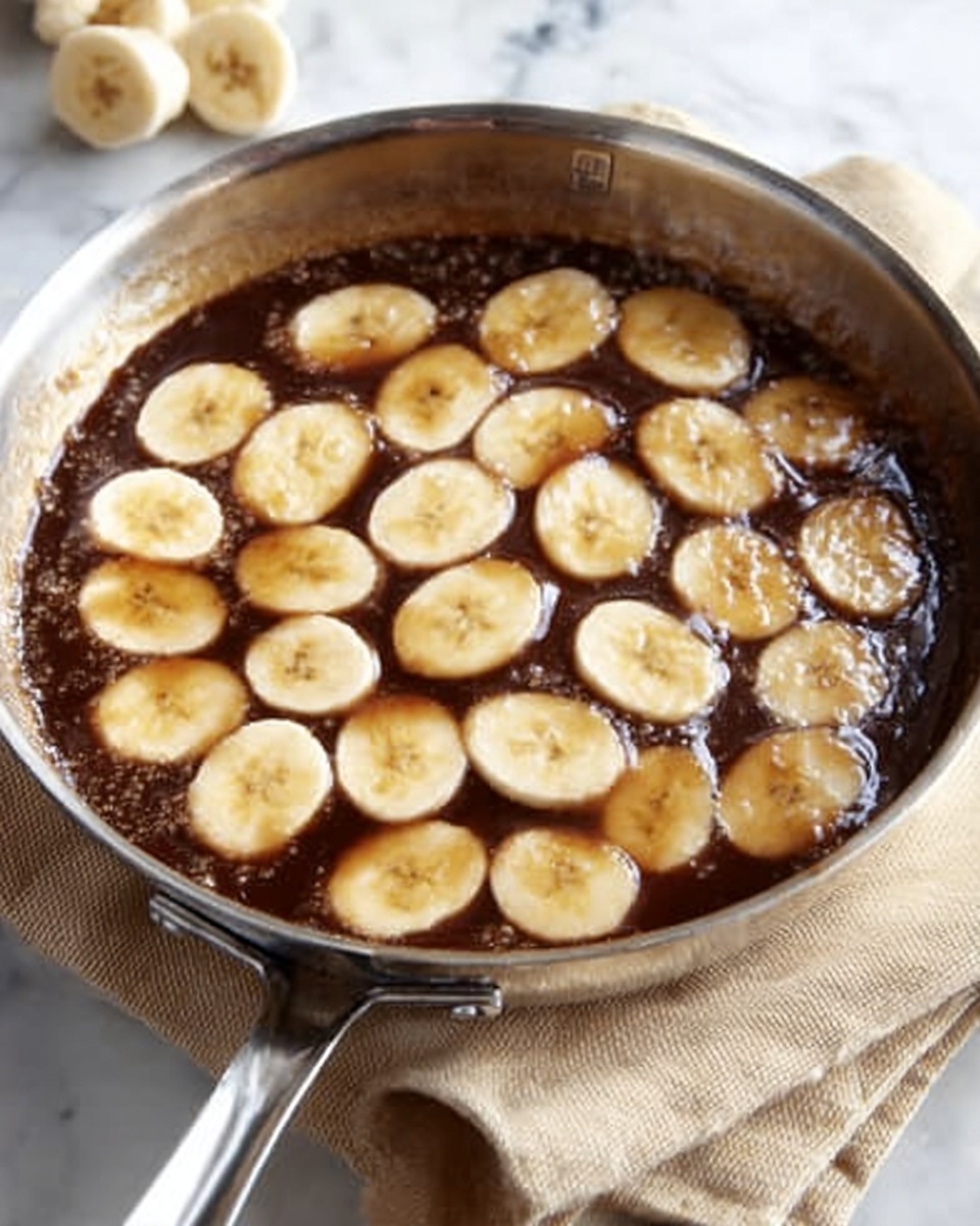 A shiny silver pan holds a dark brown thick sauce that fills the bottom evenly. Spread across the sauce are slices of banana arranged in loose rows, each slice pale yellow with a smooth texture, partially covered in the sauce which gives them a glossy shine. The pan sits on a piece of beige cloth over a white marbled surface. The edges of the pan are clean and two small handles are visible. Photo taken with an iphone --ar 4:5 --v 7