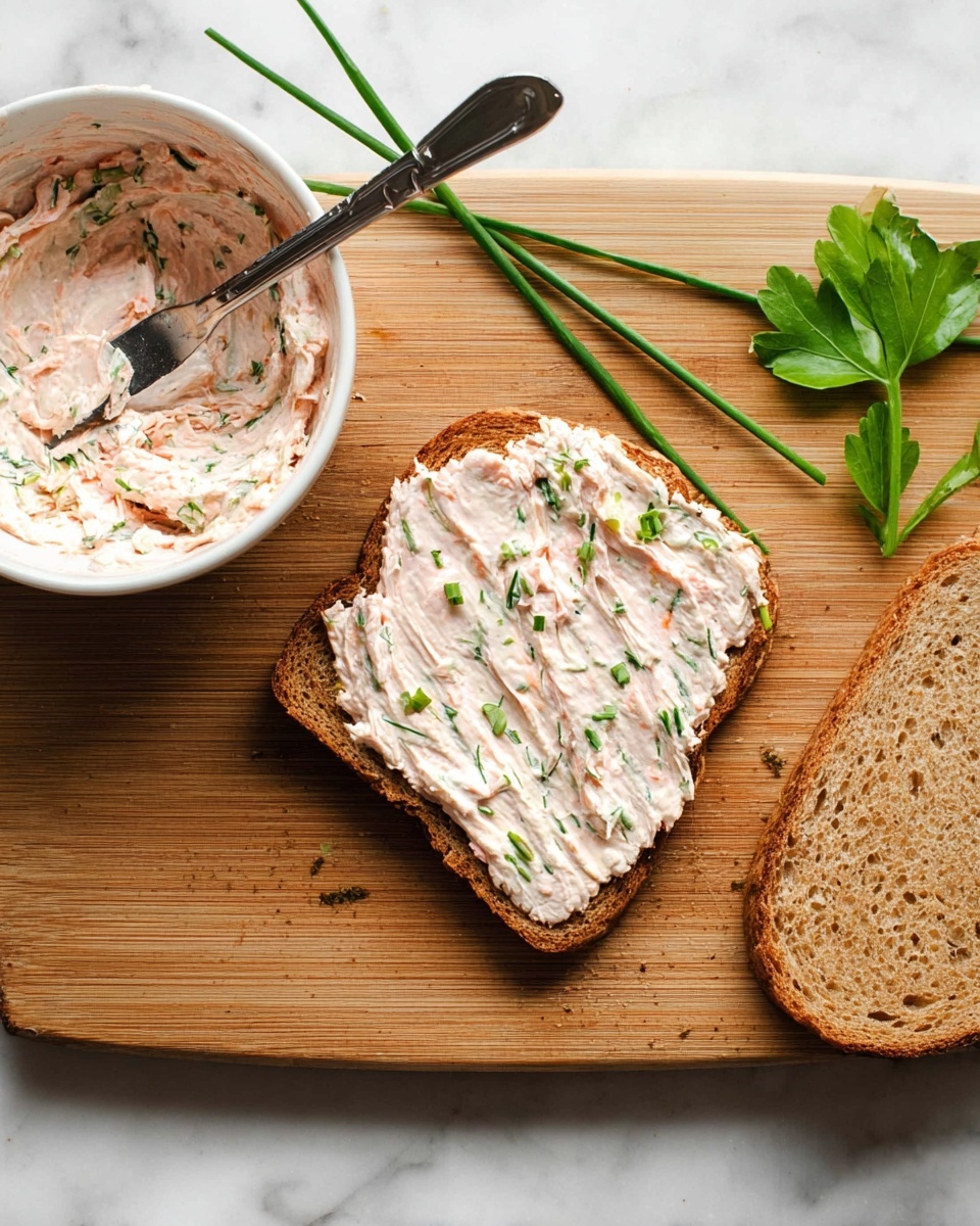 The image shows two slices of brown toasted bread on a wooden board with a white marbled background. One slice lies flat with a thick, creamy spread covering it; the spread is light pink with green herb bits mixed in, giving it a textured look. On top of this spread sits a shiny spreading knife resting diagonally. Next to the spread slice is the plain toasted bread slice. To the left side of the board is a white bowl filled with more of the same pink herb spread, and some fresh green chive stems and a parsley leaf lie near the bowl on the board. Photo taken with an iphone --ar 4:5 --v 7