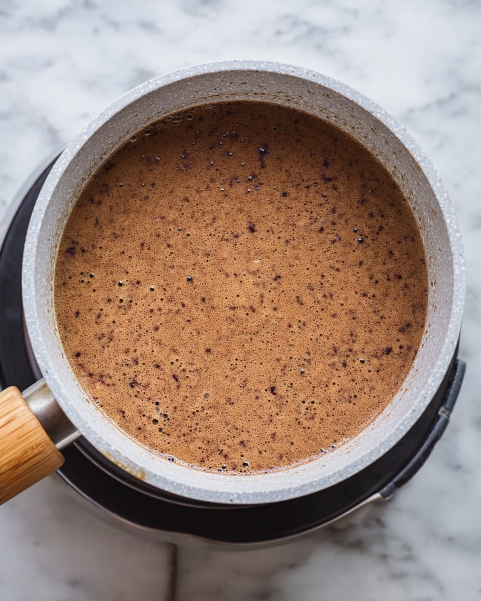 A light gray pot with a speckled texture and a wooden handle is filled with a thick brown liquid that has small black and lighter brown specks spread evenly throughout. The pot sits on a black stove burner, against a white marbled surface background. The liquid looks creamy and smooth with tiny bubbles near the edges. photo taken with an iphone --ar 4:5 --v 7