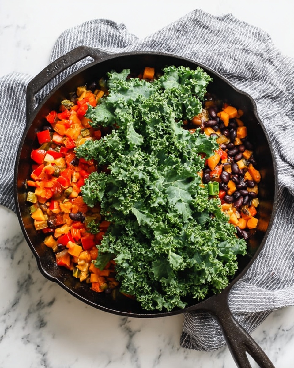 A black cast iron pan filled with a mix of colorful cooked diced vegetables and beans on the right side, including orange carrots, red bell peppers, and black beans, with large fresh green kale leaves covering the left side on top. The pan rests on a white marbled surface next to a gray and white striped cloth. Photo taken with an iphone --ar 4:5 --v 7