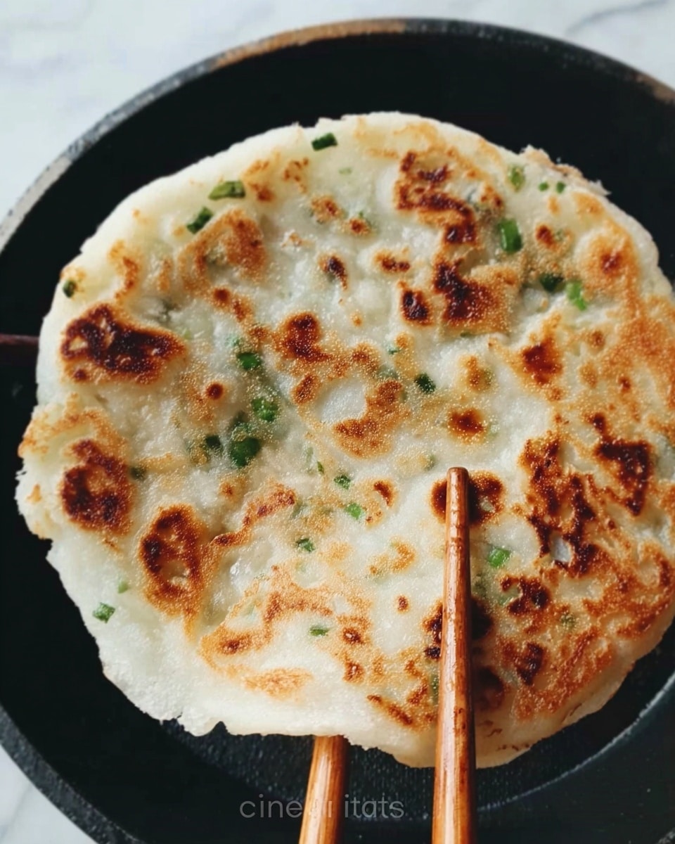 A round flat pancake is held by a pair of chopsticks over a black pan. The pancake has a golden-brown crispy surface with scattered darker brown spots, and small green pieces like chopped scallions are visible inside the translucent white dough. The edges of the pancake are slightly uneven, showing its handmade texture. The background surface is white marble. Photo taken with an iphone --ar 4:5 --v 7
