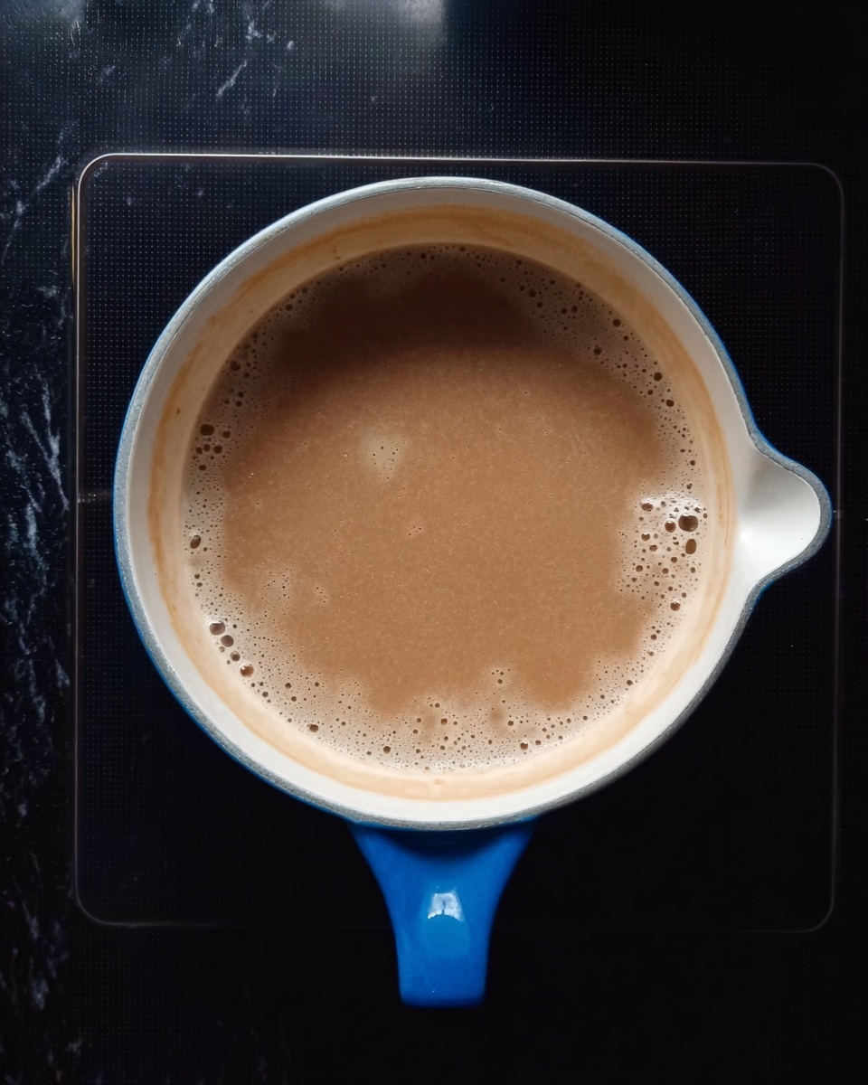 A white pan with a blue handle is on a black stove surface. Inside the pan, there is a light brown liquid with small bubbles forming on the surface. The pan is round with a small spout on one side. The background surface around the stove is a white marbled texture. Photo taken with an iphone --ar 4:5 --v 7