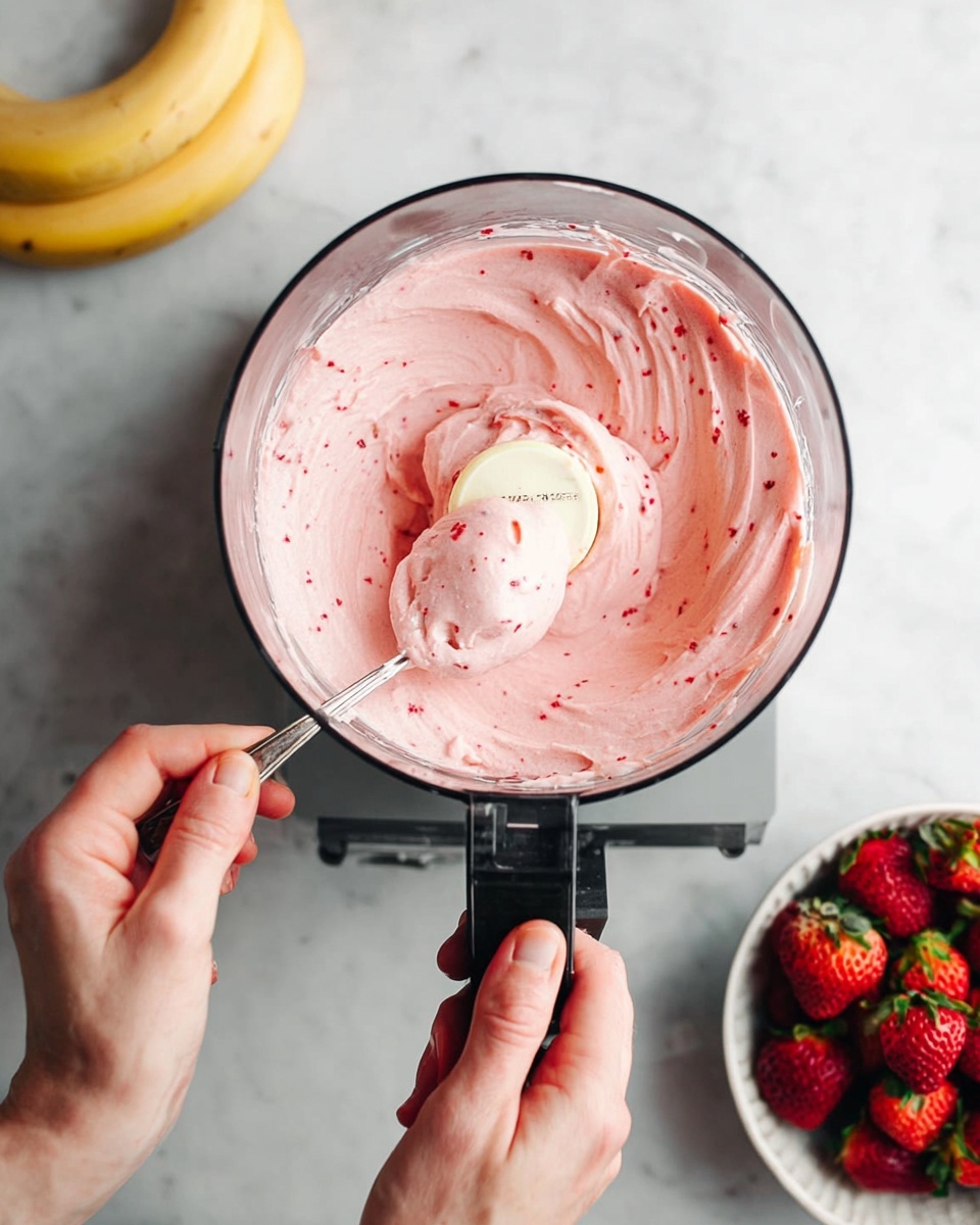 The image shows a food processor bowl filled with smooth light pink frozen dessert that has small red specks in it, swirled to form soft peaks and creamy texture on top. A woman's hand is holding a spoon scooping the mixture from the side, while the other woman's hand steadies the food processor on a white marbled surface. To the lower right side, a white bowl contains whole bright red strawberries with green leaves, and just out of frame a couple of yellow bananas are visible. The scene is bright with soft natural light coming from above. photo taken with an iphone --ar 4:5 --v 7