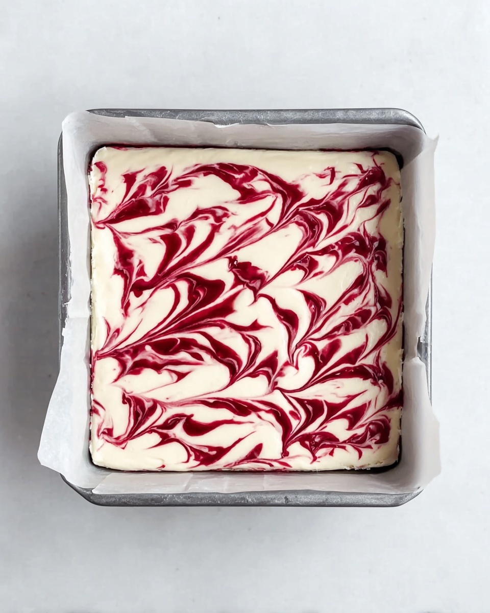A square cake pan lined with white parchment paper holds a dessert that has a thick, smooth white cream layer spread evenly on top. Swirled in the white cream are deep red streaks creating a feather-like pattern that runs horizontally across the surface. The pan is placed on a white marbled surface which adds a clean and light background to the image. photo taken with an iphone --ar 4:5 --v 7