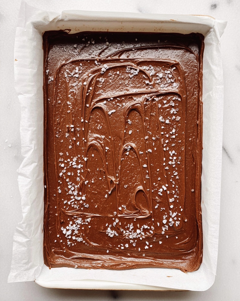 A rectangular white baking tray lined with white parchment paper holds a single thick layer of smooth, rich chocolate fudge spread evenly but with visible swirling patterns on the top. The glossy chocolate surface is sprinkled lightly with coarse flakes of sea salt, giving a contrast between the dark brown and the white salt. The tray sits on a white marbled surface. Photo taken with an iphone --ar 4:5 --v 7