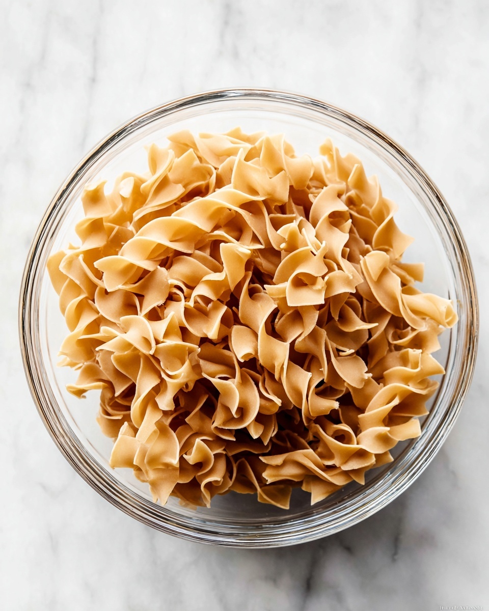A clear glass bowl filled with light brown, dried egg noodles that have ruffled edges and a slightly twisted shape, sitting on a white marbled surface. The noodles fill the bowl almost to the top, showing their smooth and firm texture with a subtle shine. The clear bowl allows you to see the noodles from the side and top, creating a layered look of overlapping noodle pieces in warm beige tones. The scene is bright, clean, and simple with no other objects around. photo taken with an iphone --ar 4:5 --v 7
