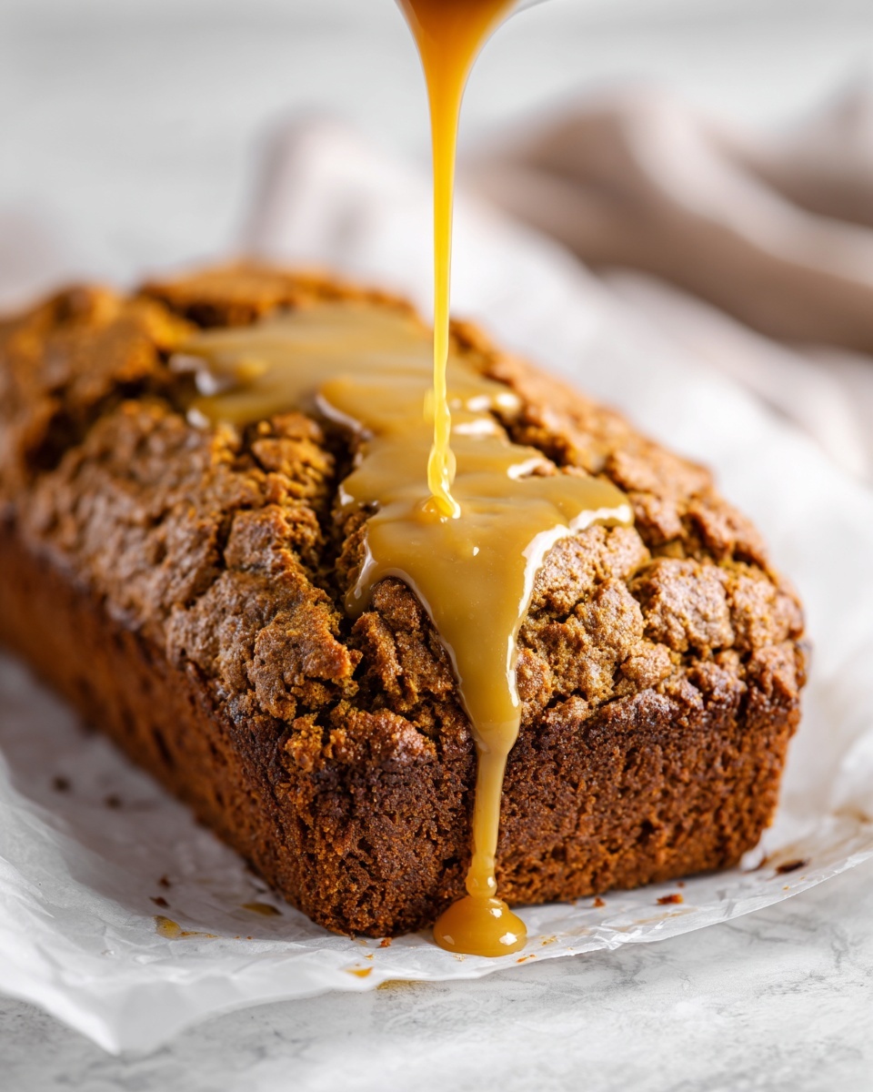A close-up of a loaf of brown pumpkin bread with a rough cracked top on white parchment paper, where a thick light brown sauce is being poured down onto the center, slowly flowing and pooling on the bread’s surface. The edges show a dense texture with darker baked sides and lighter cracks on the top. The background is a white marbled texture. photo taken with an iphone --ar 4:5 --v 7