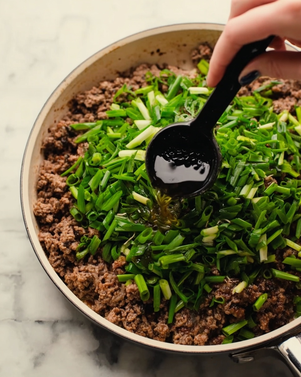 In a white pan, cooked ground meat forms the base layer with a crumbly, brown texture spread evenly. On top of the meat, there is a thick layer of fresh, chopped green onions, bright and vibrant with a crisp texture. A woman's hand holds a small black measuring spoon, pouring a clear liquid over the green onions and ground meat mixture. The background shows a white marbled surface. photo taken with an iphone --ar 4:5 --v 7