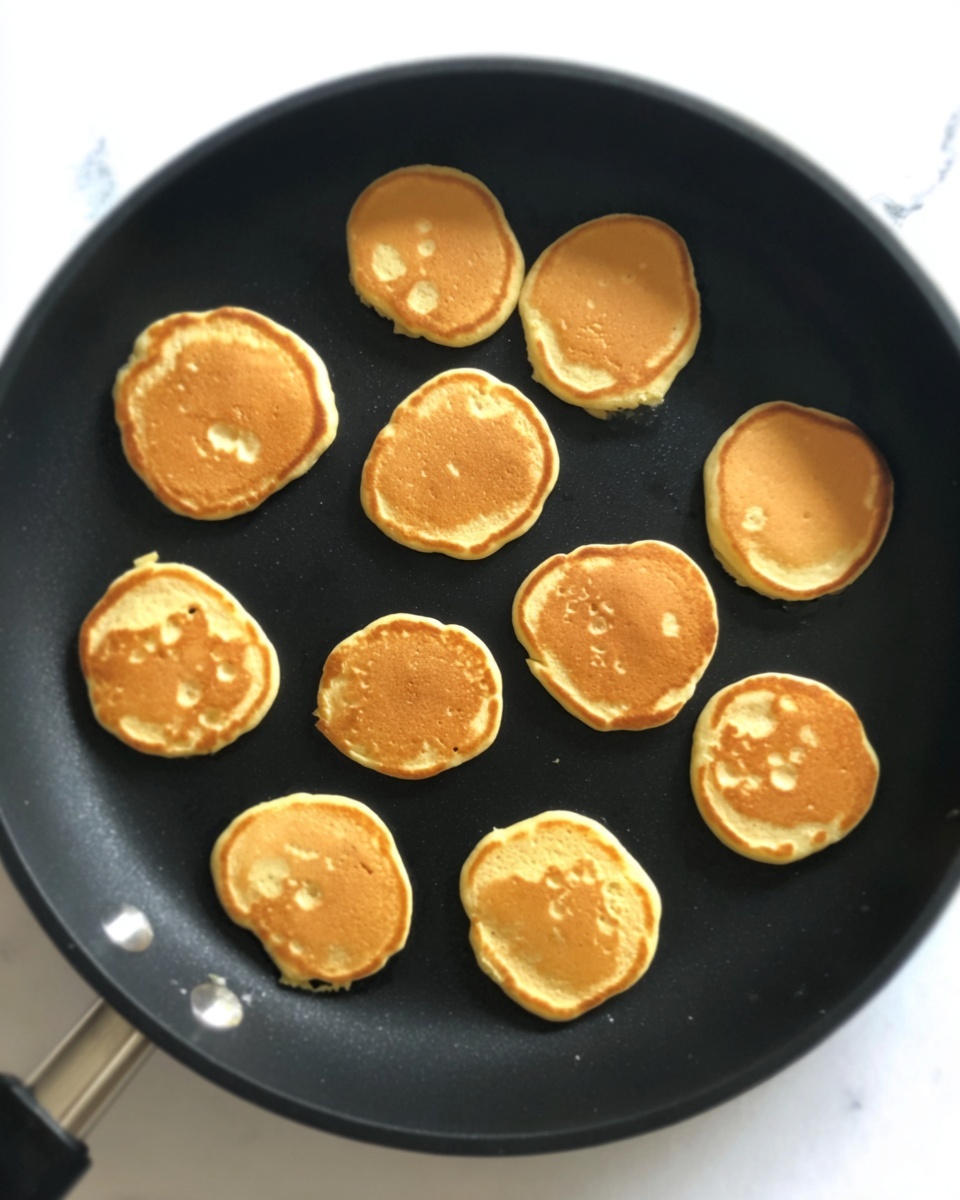 A black frying pan with twelve small, round, light brown pancakes cooking inside. The pancakes have an even golden color with some small bubbles on the surface and slightly uneven edges. The pan is set on a white marbled surface. photo taken with an iphone --ar 4:5 --v 7