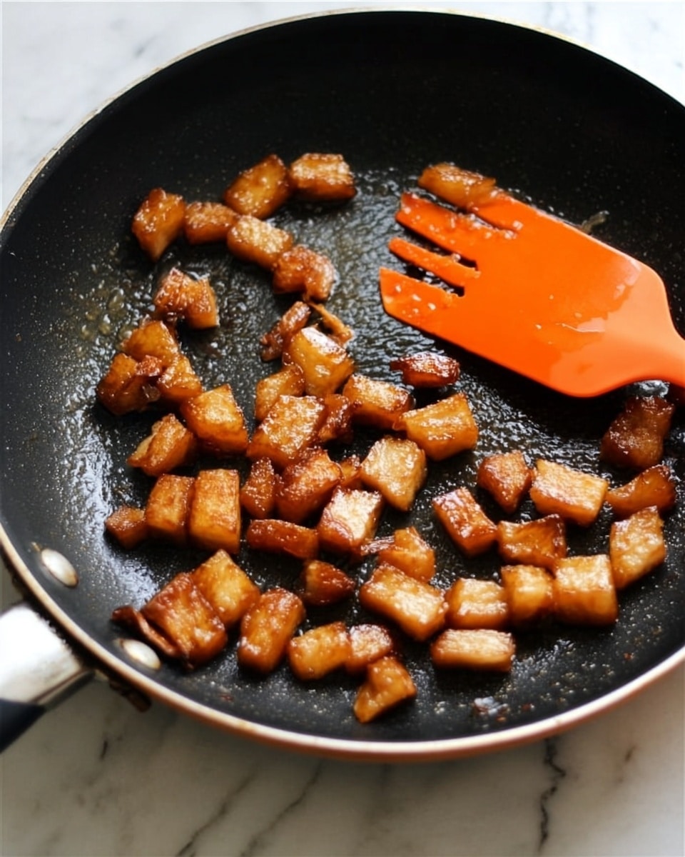 The image shows small pieces of golden-brown cooked food in a black non-stick pan. The pieces are irregular and shiny with a slightly caramelized texture. There is a bright orange spatula resting inside the pan on the right side. The background surface is white with a marbled texture. Photo taken with an iphone --ar 4:5 --v 7