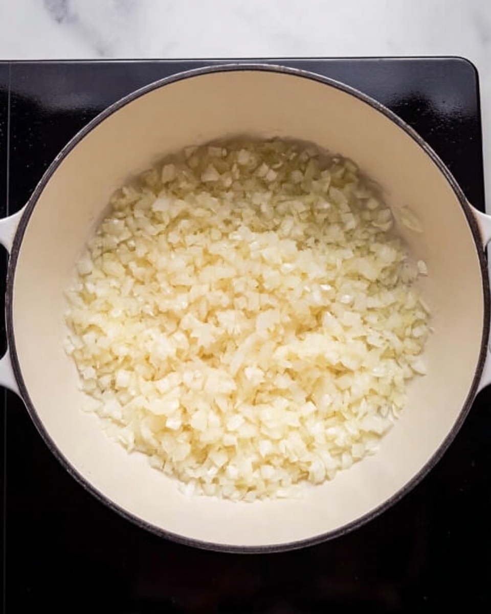 A white pot sits on a stovetop with finely chopped onions inside. The onions are pale yellow and white, spread evenly across the bottom of the pot. The pot is round and deep, showing a smooth texture inside, with a shiny black stovetop under it. The background has a clean white marbled surface. photo taken with an iphone --ar 4:5 --v 7