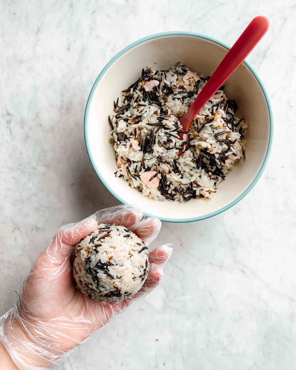 A white bowl with a light blue rim is filled with a mixed rice dish, showing white rice grains combined with dark seaweed flakes and small pieces of light pink fish, with a red spoon resting inside the bowl. Next to it, a woman's hand wearing a clear plastic glove holds a round rice ball made from the same mixture, showing visible layers of white rice, dark seaweed, and pink fish pieces evenly spread throughout. The background and surface are a white marbled texture. photo taken with an iphone --ar 4:5 --v 7
