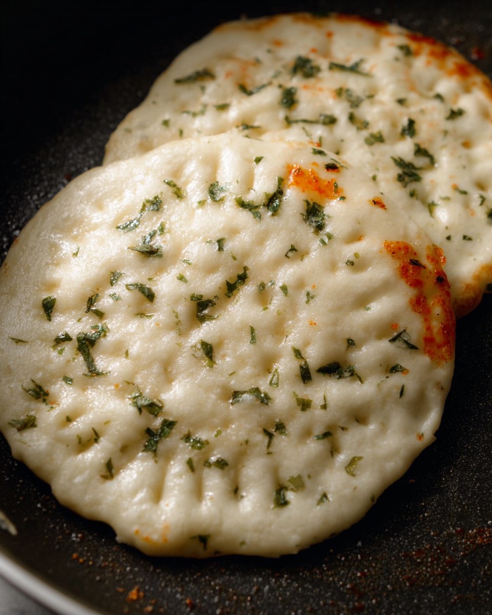 Two flat, round dough pieces cooked in a pan, each about one layer thick with a slightly puffy, uneven white surface. The dough has small bubbles and is dotted with tiny green herb flakes scattered across the top. The edges are pressed down with a pattern of small fork marks, showing some slight orange-red sauce peeking out from underneath near the edges. The pan is dark and contrasts with the light dough, making the texture and herb details clear. photo taken with an iphone --ar 4:5 --v 7