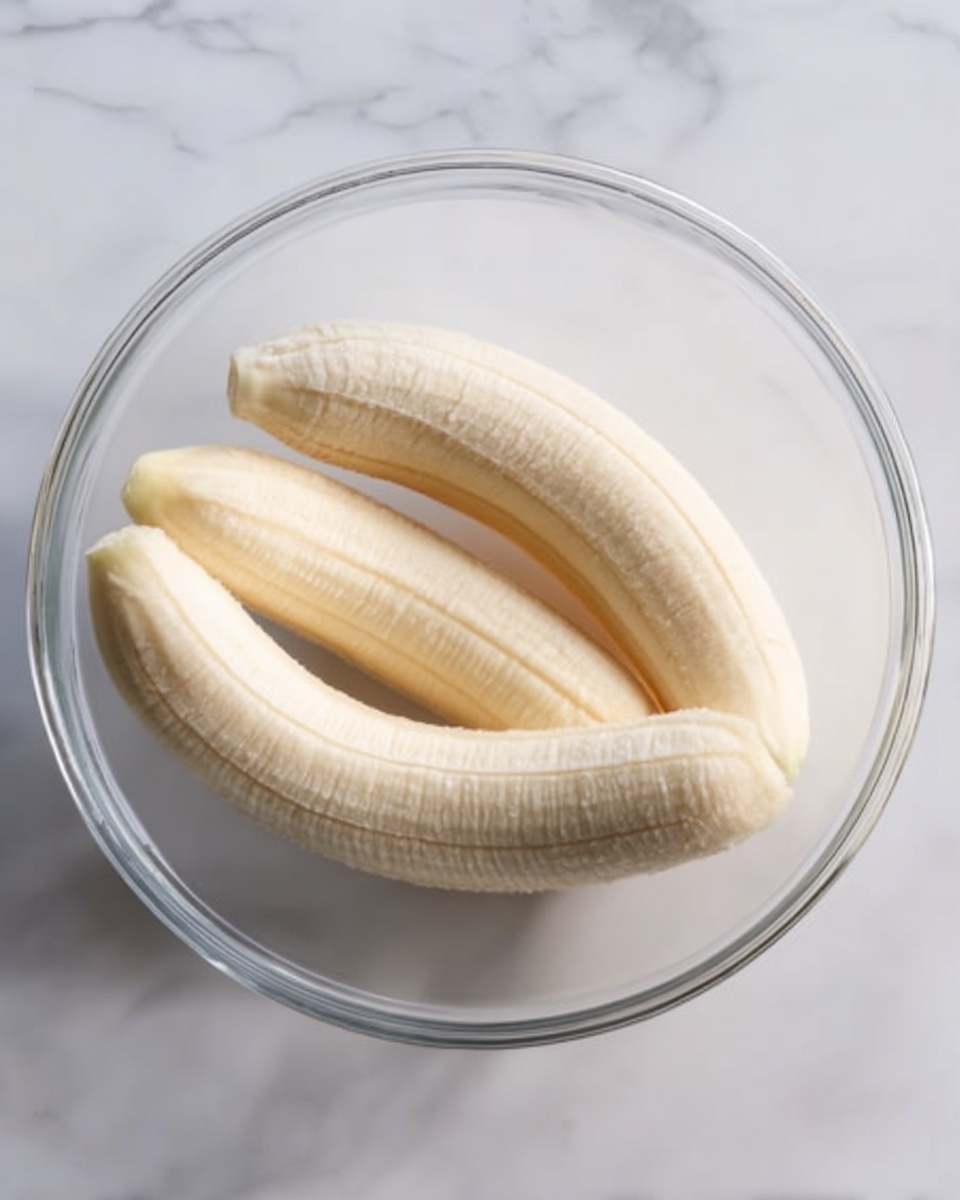 In the image, there is a clear glass bowl sitting on a white marbled surface. Inside the bowl, three whole peeled bananas rest close together. The bananas are smooth and pale yellow with visible ridges. The bowl has a simple round shape with transparent sides. The lighting softly highlights the texture of the bananas and the clear glass, creating subtle shadows within the bowl. Photo taken with an iphone --ar 4:5 --v 7