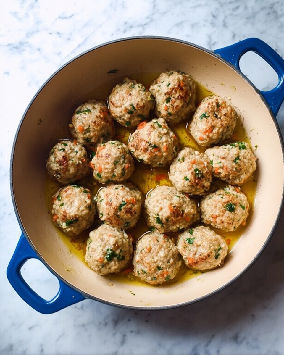 A round beige pan with blue handles filled with about fifteen light brown meatballs with bits of green herbs and orange carrots throughout. The meatballs are evenly spaced in the pan on a shallow pool of golden cooking oil. The pan sits on a white marbled surface. photo taken with an iphone --ar 4:5 --v 7