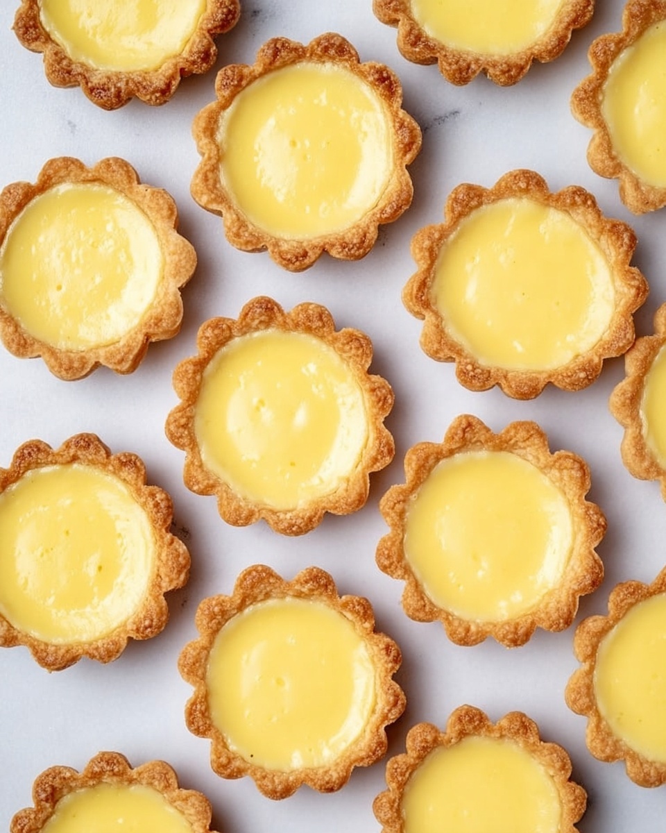 The image shows many small tartlets arranged on a white marbled surface. Each tartlet has a golden brown crust with scalloped edges and is filled with a smooth, creamy yellow filling that looks slightly shiny. The filling sits evenly inside the crust, giving each tartlet a neat, round shape. The tartlets are closely placed but not touching each other, filling the frame with their warm colors and soft textures. Photo taken with an iphone --ar 4:5 --v 7