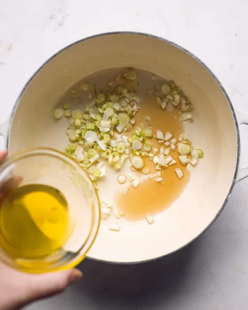 A white pot sits on a white marbled surface, inside which there are small pieces of white and light green chopped onions spread around light brown liquid, possibly oil or sauce. A woman's hand holding a glass bowl with a yellow liquid or ingredient is seen in the lower right corner, about to pour it into the pot. The scene is simple and focused on the beginning of cooking with soft, neutral colors. Photo taken with an iphone --ar 4:5 --v 7