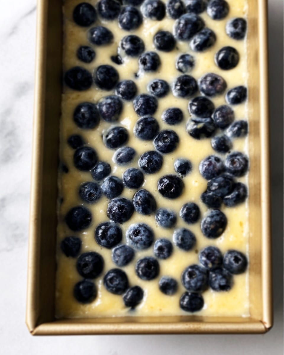 The image shows a rectangular cake pan filled with light yellow batter dotted generously with whole fresh blueberries spread evenly across the top layer. The batter has a smooth, slightly shiny texture with the dark blue blueberries creating a strong color contrast. The pan edge is metal and the background is a white marbled surface. Photo taken with an iphone --ar 4:5 --v 7