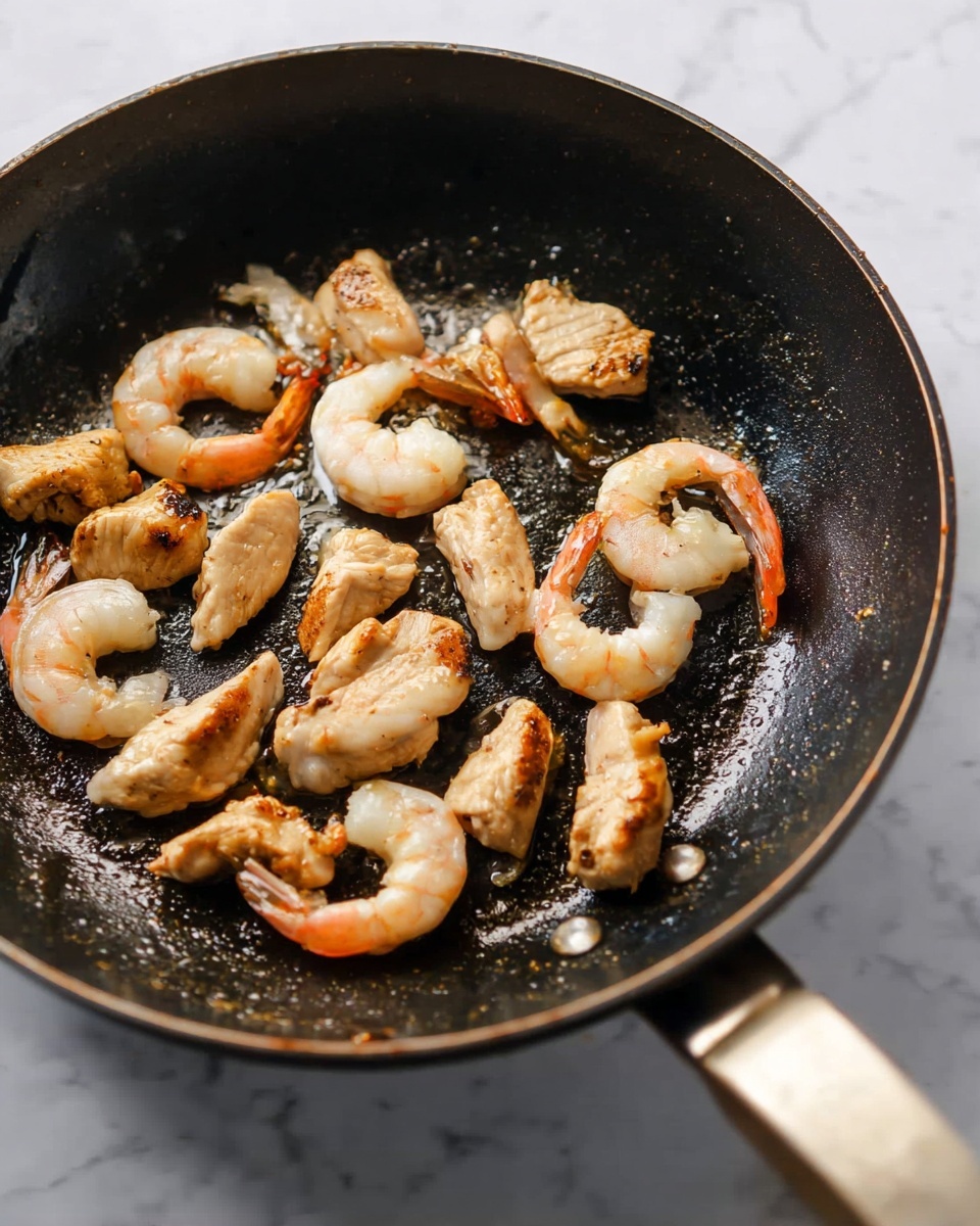 Inside a dark black pan with a shiny surface, there are several shrimp and pieces of light golden brown chicken cooking. The shrimp are curved with pinkish tails and a slightly translucent white and light pink body. The chicken pieces are small and irregular shaped with a seared outer layer that is golden brown. The pan handle is visible on the right side with a light brown grip. The background is a white marbled texture. photo taken with an iphone --ar 4:5 --v 7
