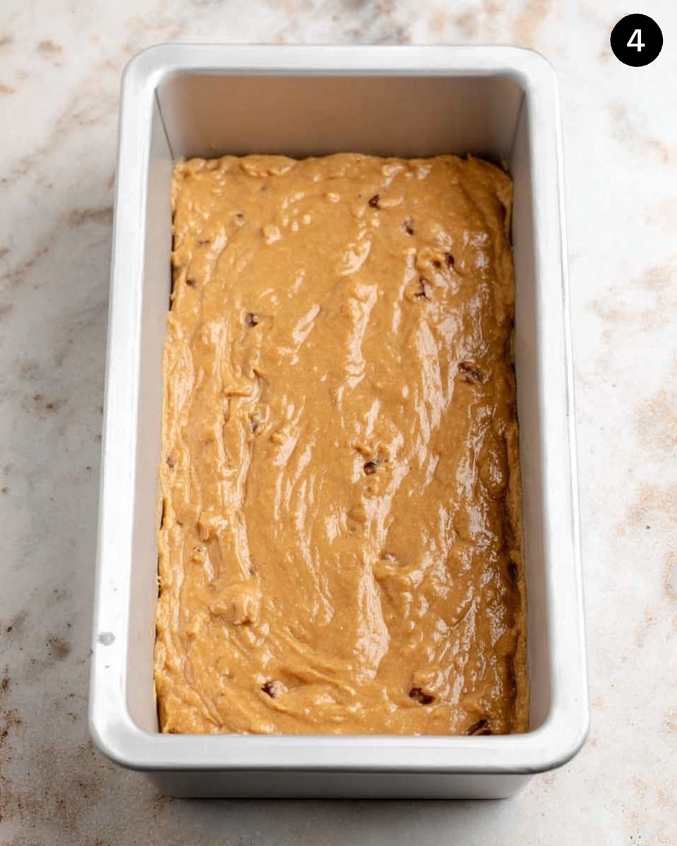 A rectangular white metal baking pan filled with a thick, light brown batter with small chunks visible throughout. The batter is spread evenly inside the pan, which is placed on a white marbled surface. The texture of the batter looks smooth with some small lumps, showing it is ready to be baked. Photo taken with an iphone --ar 4:5 --v 7
