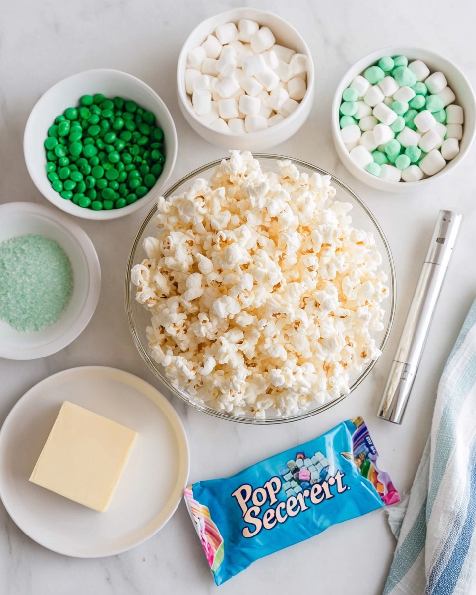 The image shows a top view of several small white bowls and a clear glass bowl arranged on a white marbled surface. The clear glass bowl in the center is filled with light, fluffy popcorn with a slight yellow tint. Surrounding it are white bowls with different ingredients: one bowl is full of small white mini marshmallows, one has round green and white candies, and another holds green sugar crystals. Next to these is a small white plate with a rectangular piece of butter. There is also a colorful packet labeled