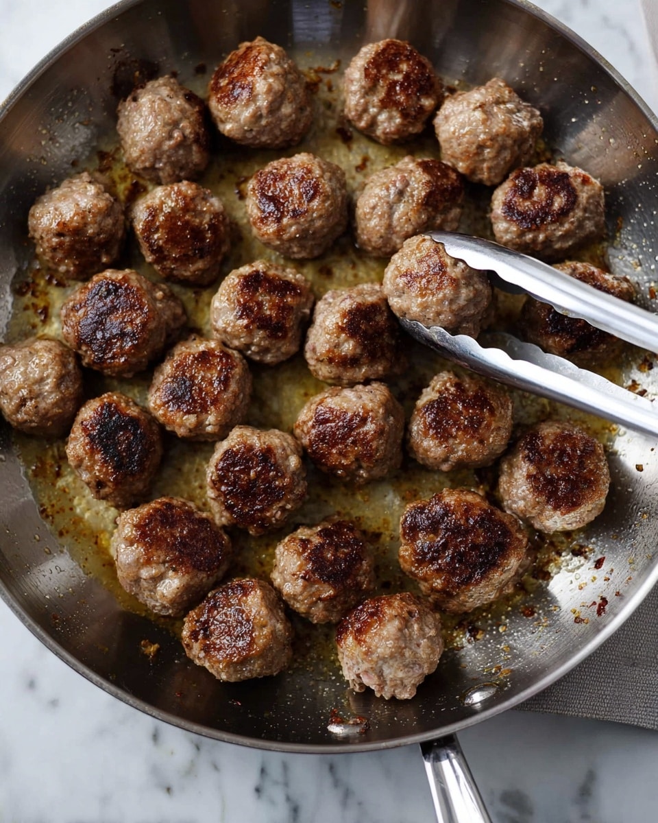 A silver pan filled with about twenty browned meatballs, each roughly round with a slightly rough texture showing browned crust spots and some areas of lighter cooked meat, resting evenly across the pan's bottom. A pair of silver tongs holds one meatball near the right side of the pan. The pan's surface inside shows small browned bits and light reflections from cooking oil. The background is a white marbled texture. photo taken with an iphone --ar 4:5 --v 7