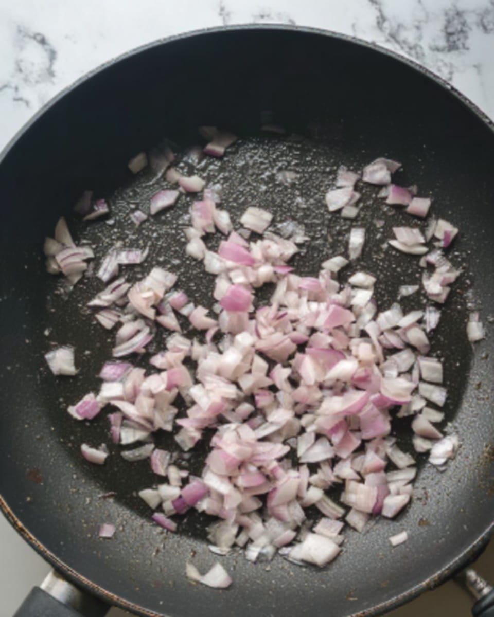A close-up of a black frying pan on a white marbled surface with small, pale pink and white pieces of chopped shallots cooking in it. The shallots are spread evenly in one layer, showing a glossy texture as they soften. A woman's hand is visible at the edge of the pan, near the top left corner, holding or stirring them. The black pan contrasts with the light shallots and the bright white marble background, highlighting the cooking process. photo taken with an iphone --ar 4:5 --v 7