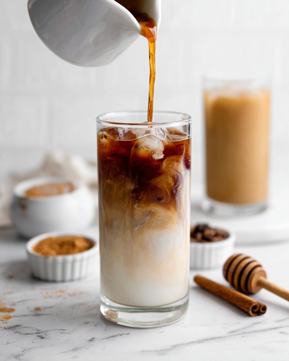 A tall clear glass filled mostly with white milk and ice cubes at the bottom and middle, with a layer of dark brown coffee being poured from a white small pitcher into the top, creating a swirling mix where the coffee meets the milk. Behind it, there is a second glass with a fully mixed light brown iced coffee. The setup is on a white marbled surface with small white ramekins, one holding a wooden honey dipper, and some cinnamon sticks scattered nearby. The background is simple and white. photo taken with an iphone --ar 4:5 --v 7