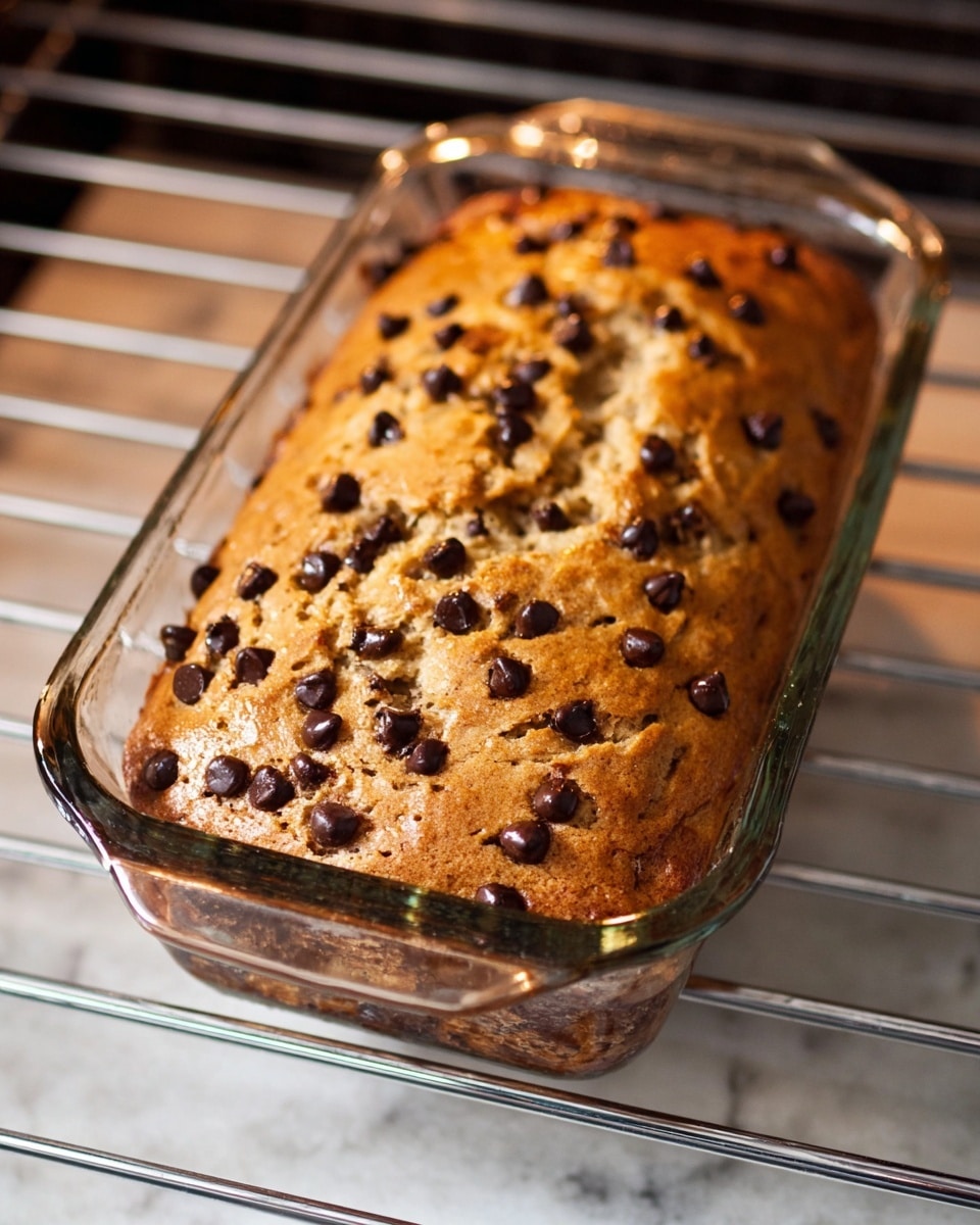 A glass loaf pan holds a golden-brown baked banana bread studded with numerous dark brown chocolate chips spread evenly on the top and throughout the visible sides, sitting on a silver oven rack inside the oven. The bread has a slightly cracked, rough texture on the top surface with a soft, moist look inside. The clear glass pan shows the thickness and the even baking of the bread, resting on a white marbled texture. photo taken with an iphone --ar 4:5 --v 7