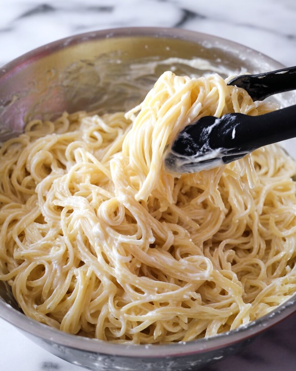 The image shows a close-up of a large silver metal bowl filled with cooked spaghetti noodles coated in a creamy, light beige sauce. The noodles have a soft and slightly sticky texture, swirling in thick layers filling the bowl. A black kitchen tong is lifting a portion of the spaghetti strands from the bowl, highlighting the glossy sauce that clings to the noodles. The background surface has a white marbled texture. photo taken with an iphone --ar 4:5 --v 7