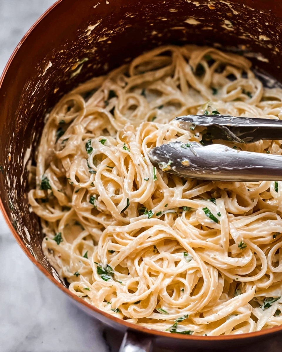 The image shows a close-up of creamy pasta mixed with green herbs inside a brown cooking pot. The pasta is light beige with a smooth, creamy texture, while the small green herb pieces add contrast throughout. A pair of silver tongs is partially visible, holding the pasta and slightly covered with the creamy sauce. The pot's inside surface has some sauce splattered on it. The close view highlights the tangled, soft noodles and the creamy sauce coating each strand. The image background features a white marbled texture. Photo taken with an iphone --ar 4:5 --v 7