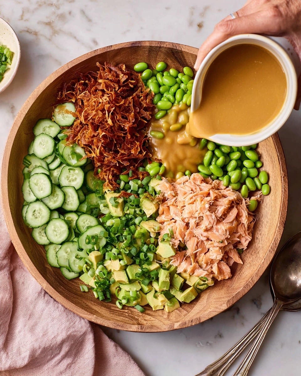 A large wooden bowl holds a colorful salad with six distinct layers arranged in sections: thinly sliced green cucumbers on the left, crispy browned shredded topping at the top, bright green edamame beans in the middle, diced light green avocado on the right, and flaked light pink cooked salmon at the bottom. Chopped green onions are sprinkled over the salmon and edamame. A woman's hand pours a thick, tan sauce from a small white bowl over the salad. The bowl sits on a white marbled surface next to a light pink cloth and a silver spoon. Photo taken with an iphone --ar 4:5 --v 7