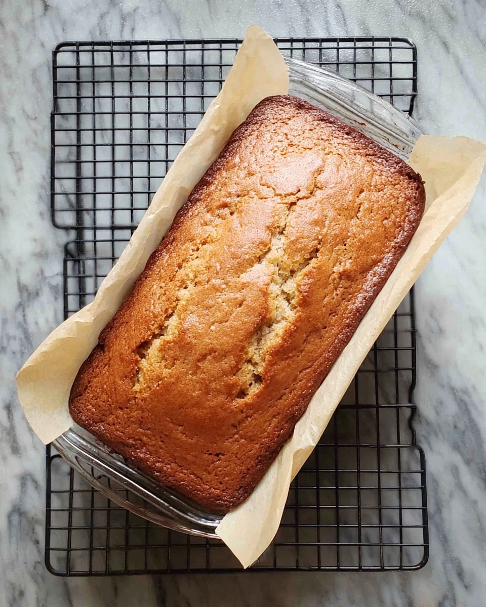 A golden-brown loaf cake with a slightly cracked top layer, showing a soft and moist inside texture beneath. It is baked in a clear glass rectangular dish lined with off-white parchment paper that folds over the edges, resting on a black cooling rack with a grid pattern. The whole setup is placed on a white marbled surface. photo taken with an iphone --ar 4:5 --v 7