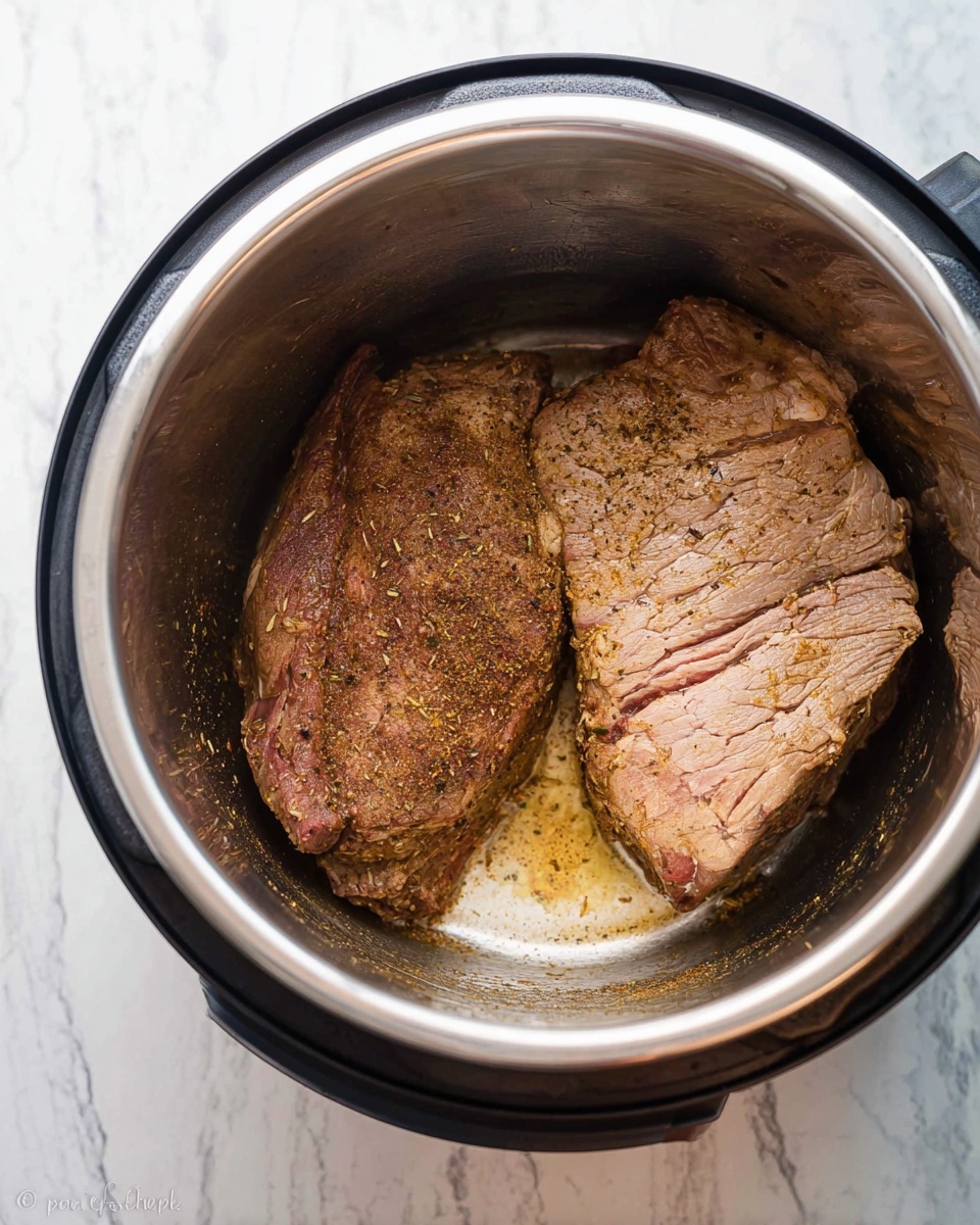 Inside a shiny silver pressure cooker, there are two large pieces of browned meat resting close together. The meat has a rough texture with visible seasoning in brown and green specks spread over its surface. The larger piece on the right shows folds and creases in a light brown color with some pinkish areas. The smaller piece on the left is darker brown with a firmer texture. The bottom of the cooker has some browned bits and a small pool of light brown oil. The background surface is a white marbled texture. photo taken with an iphone --ar 4:5 --v 7