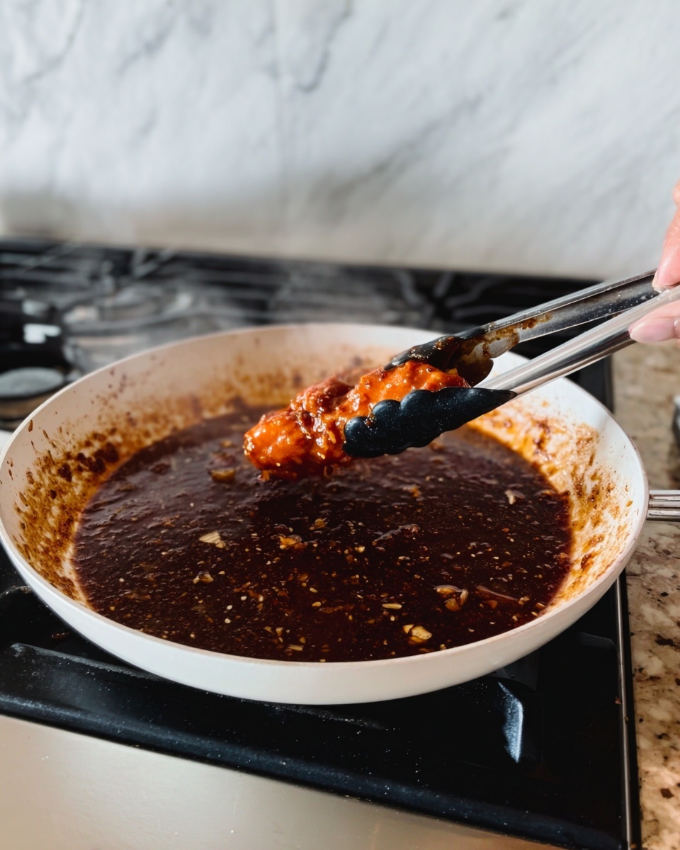 The image shows a close-up of a white pan on a white marbled stove surface filled with a dark brown sauce with small chunks. A pair of black and white tongs held by a woman's hand is lifting a bright orange piece of food coated with the sauce. The sauce looks thick and shiny with bits of garlic or spices scattered throughout. Steam rises slightly from the pan, showing the food is hot. Photo taken with an iphone --ar 4:5 --v 7